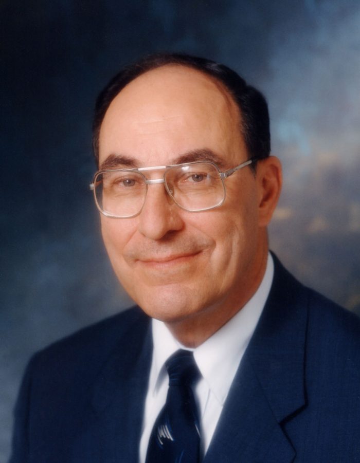 Portrait of smiling Dr Hall in blue suit, tie and wireframe glasses.